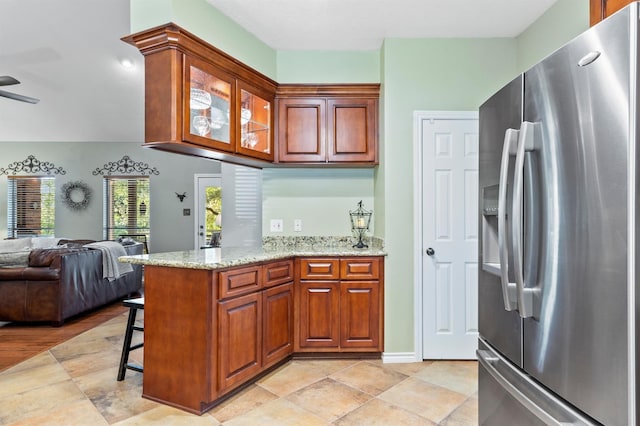 kitchen with stainless steel refrigerator with ice dispenser, kitchen peninsula, ceiling fan, light stone counters, and a breakfast bar area