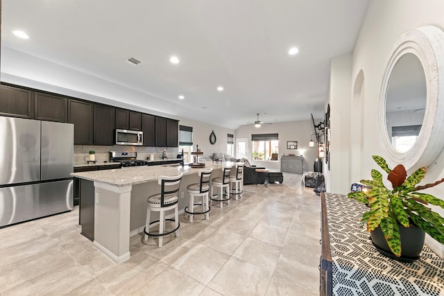 kitchen featuring stainless steel appliances, ceiling fan, light stone counters, a center island with sink, and a breakfast bar area