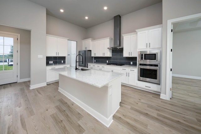 kitchen featuring light stone countertops, appliances with stainless steel finishes, wall chimney exhaust hood, white cabinetry, and an island with sink