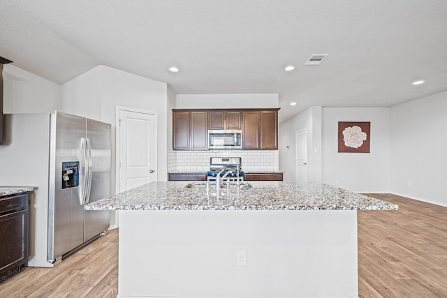 kitchen with stainless steel appliances, backsplash, a kitchen island with sink, light stone counters, and dark brown cabinets