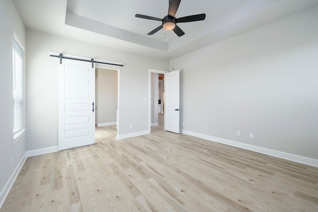 unfurnished bedroom featuring ceiling fan, a barn door, light hardwood / wood-style flooring, and a tray ceiling