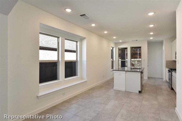 kitchen with a center island with sink, gas stove, tasteful backsplash, dark stone counters, and white cabinets