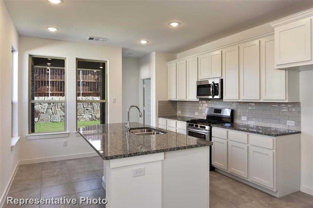 kitchen with an island with sink, appliances with stainless steel finishes, dark stone counters, white cabinets, and sink