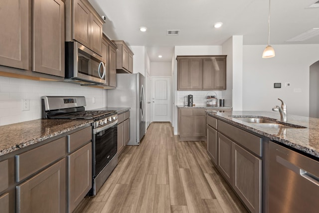 kitchen with decorative light fixtures, stainless steel appliances, dark stone counters, sink, and light hardwood / wood-style flooring