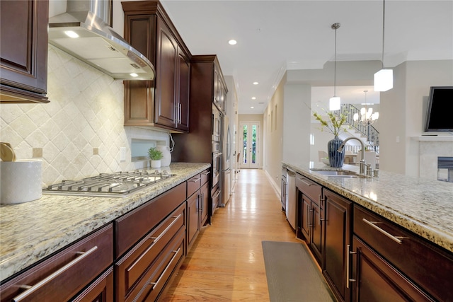 kitchen with wall chimney range hood, sink, appliances with stainless steel finishes, light stone counters, and a chandelier