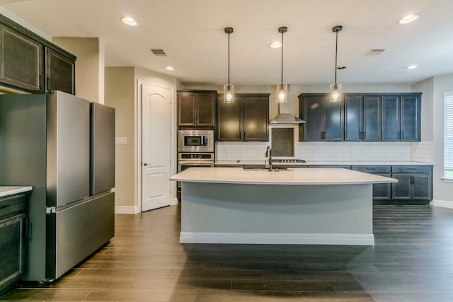 kitchen with decorative light fixtures, wall chimney range hood, stainless steel appliances, an island with sink, and backsplash