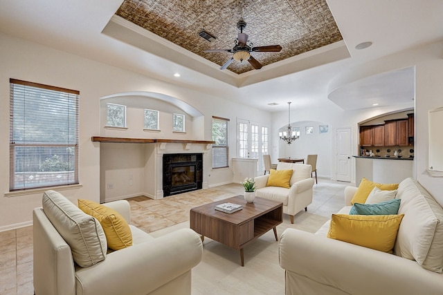 living room featuring light tile patterned flooring, ornamental molding, ceiling fan with notable chandelier, and a raised ceiling