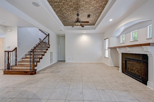 unfurnished living room featuring ceiling fan, a tray ceiling, a tiled fireplace, and light tile patterned flooring