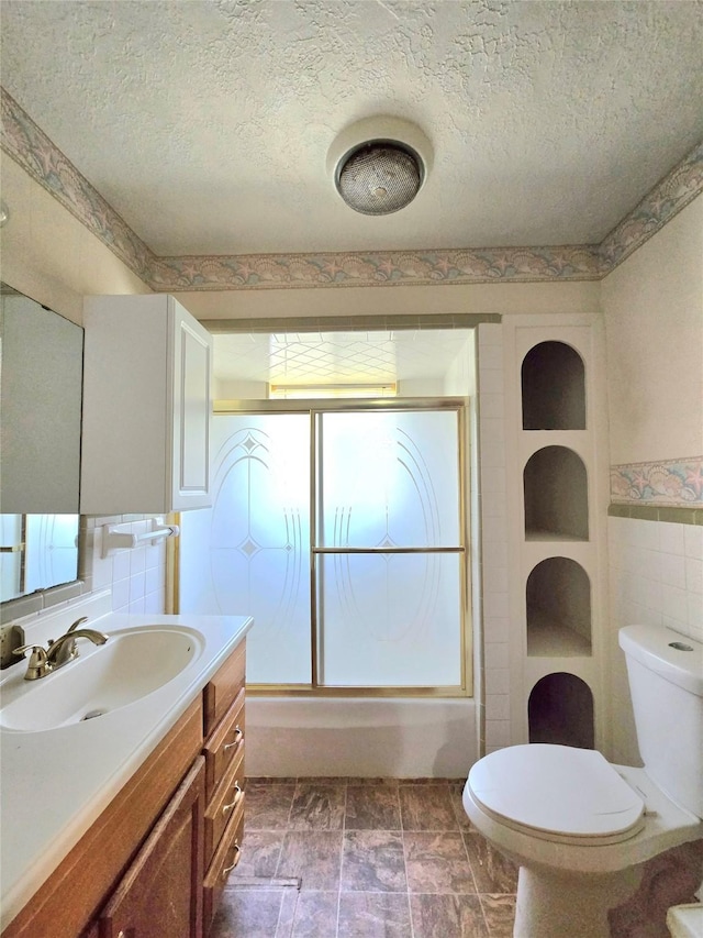 full bathroom featuring toilet, bath / shower combo with glass door, tile walls, a textured ceiling, and vanity