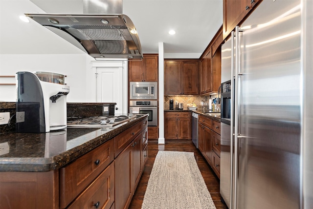 kitchen featuring backsplash, range hood, dark wood-type flooring, appliances with stainless steel finishes, and dark stone counters