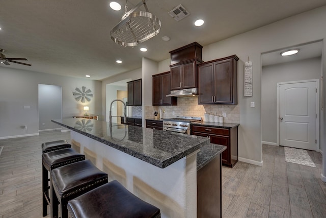 kitchen featuring high end range, light hardwood / wood-style floors, a kitchen bar, dark stone counters, and an island with sink