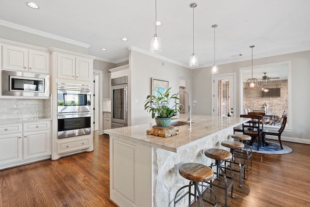 kitchen with appliances with stainless steel finishes, hanging light fixtures, ceiling fan, light stone counters, and a center island with sink