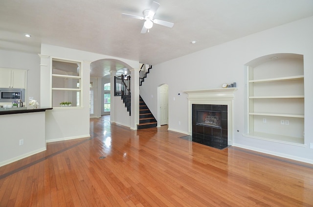 unfurnished living room with wood-type flooring, ornate columns, built in features, ceiling fan, and a tiled fireplace