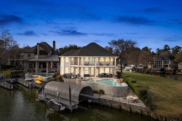 back house at dusk with a balcony, a swimming pool with hot tub, a patio area, a water view, and a lawn