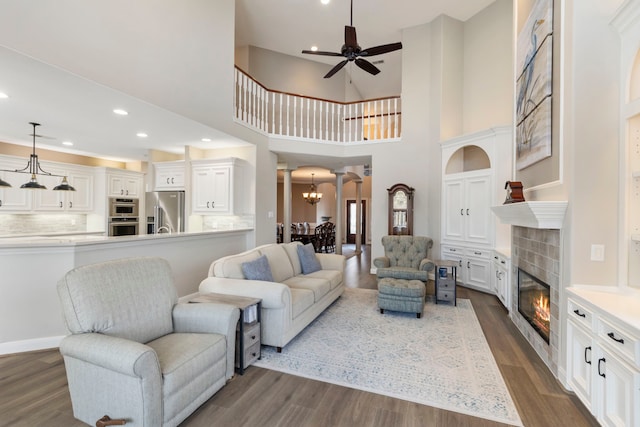 living room featuring ornate columns, dark hardwood / wood-style flooring, a tiled fireplace, a high ceiling, and ceiling fan with notable chandelier