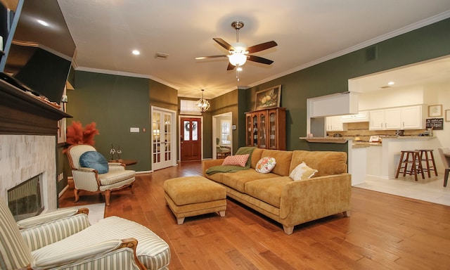 living room featuring ceiling fan with notable chandelier, french doors, light hardwood / wood-style floors, ornamental molding, and a tile fireplace