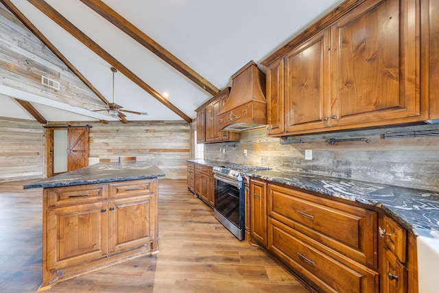 kitchen with dark stone counters, vaulted ceiling with beams, stainless steel gas range oven, premium range hood, and light wood-type flooring