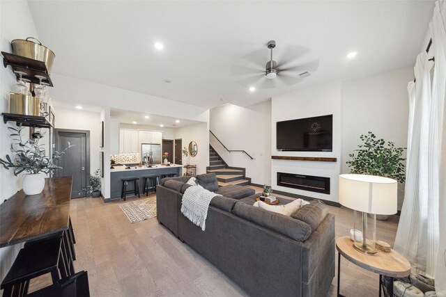 living room with ceiling fan, a glass covered fireplace, light wood-style flooring, recessed lighting, and stairway