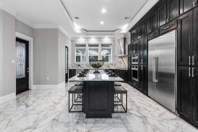 kitchen with appliances with stainless steel finishes, wall chimney exhaust hood, a kitchen island, light stone counters, and a breakfast bar area