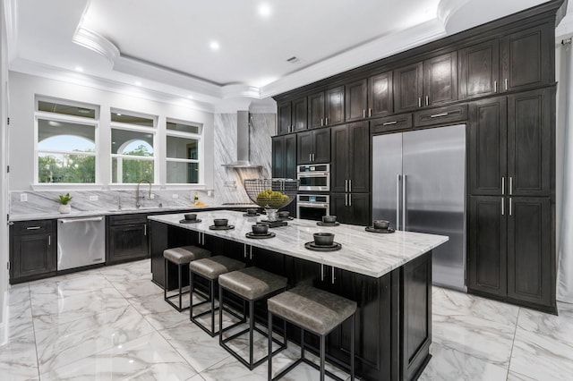 kitchen with a breakfast bar area, appliances with stainless steel finishes, a raised ceiling, wall chimney range hood, and a center island