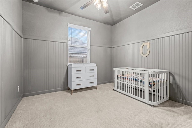 carpeted bedroom featuring a nursery area, a ceiling fan, visible vents, and wainscoting