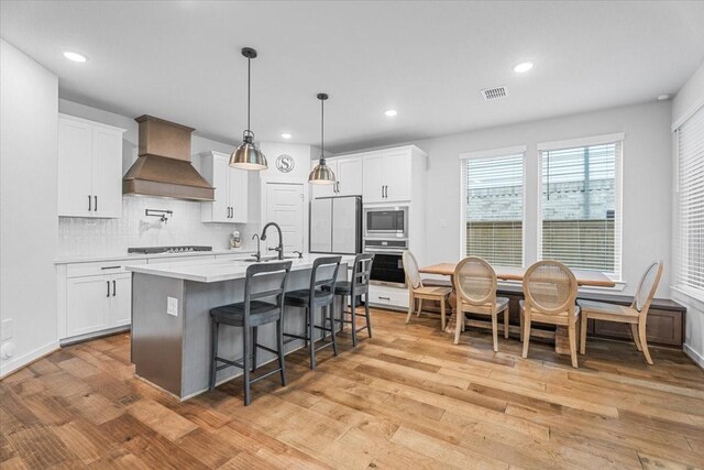 kitchen featuring custom exhaust hood, appliances with stainless steel finishes, white cabinets, sink, and a center island with sink