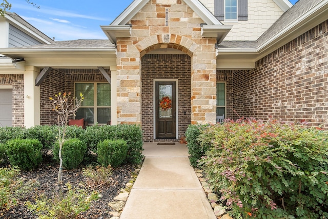 view of exterior entry with a shingled roof, a garage, stone siding, and brick siding