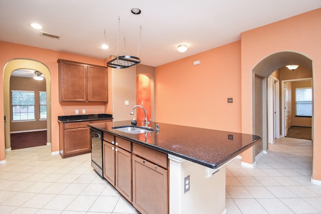 kitchen featuring sink, light tile patterned floors, an island with sink, black dishwasher, and dark stone countertops