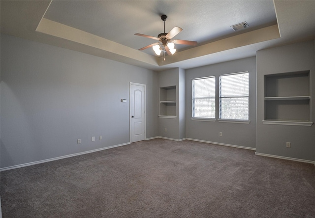 carpeted empty room with ceiling fan, built in shelves, and a raised ceiling