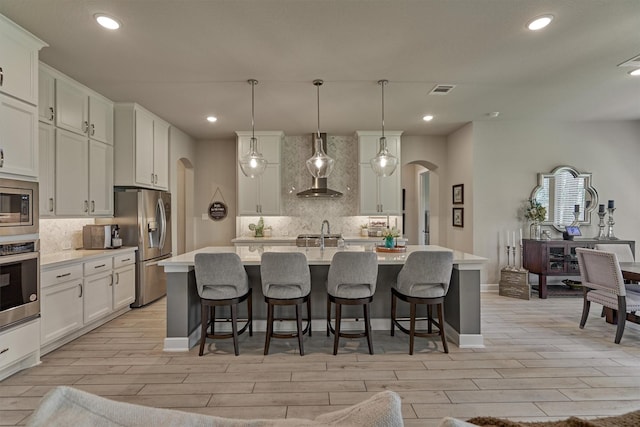 kitchen featuring decorative light fixtures, a kitchen island with sink, wall chimney range hood, and stainless steel appliances