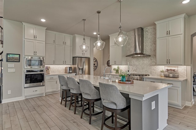 kitchen with white cabinetry, stainless steel appliances, wall chimney exhaust hood, and a center island with sink