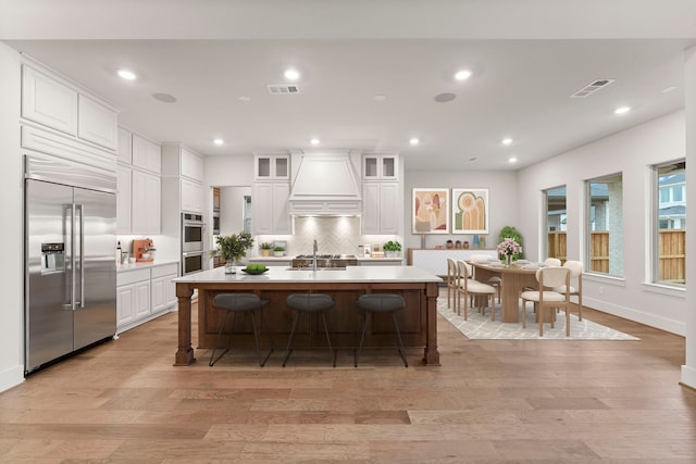 kitchen with stainless steel appliances, an island with sink, custom range hood, and white cabinets