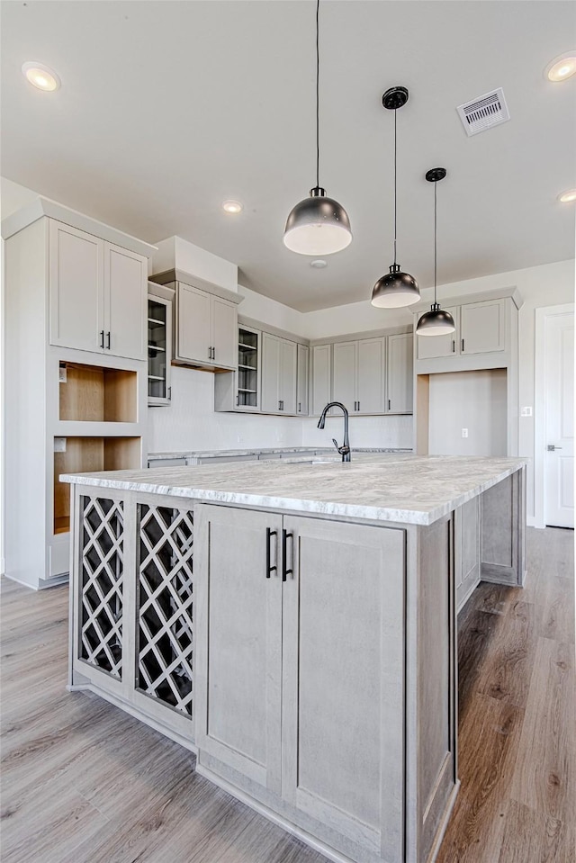 kitchen featuring light hardwood / wood-style floors, a center island with sink, gray cabinets, hanging light fixtures, and light stone countertops