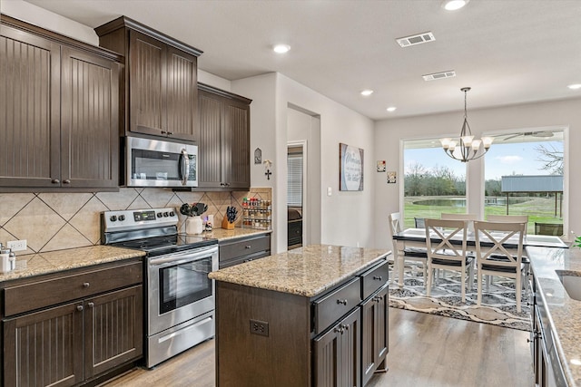 kitchen featuring dark brown cabinetry, a center island, stainless steel appliances, an inviting chandelier, and light stone counters