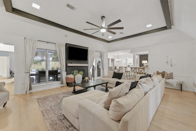 living room with ceiling fan with notable chandelier, a large fireplace, a tray ceiling, crown molding, and light wood-type flooring