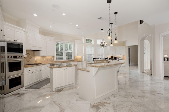 kitchen with white cabinets, stainless steel appliances, hanging light fixtures, light stone counters, and a center island with sink