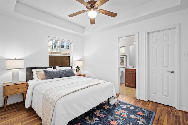 bedroom with a tray ceiling, light hardwood / wood-style flooring, ensuite bath, and ceiling fan