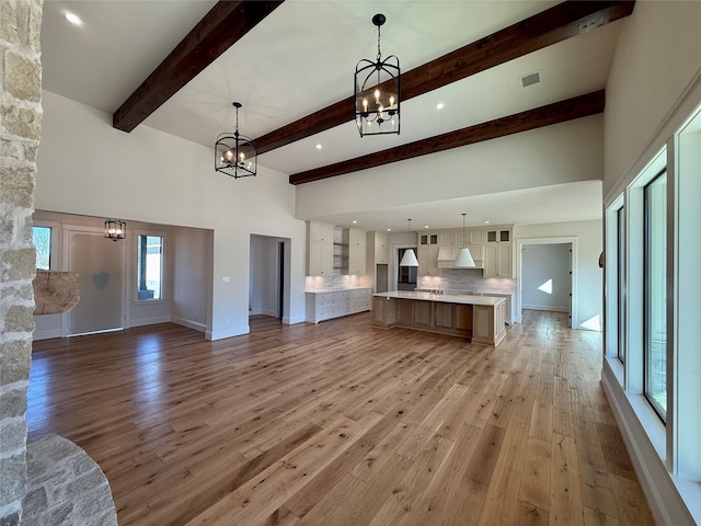 unfurnished living room featuring beam ceiling, a towering ceiling, a chandelier, and light hardwood / wood-style flooring