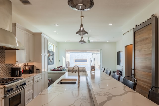 kitchen featuring high end stove, hanging light fixtures, sink, a breakfast bar, and wall chimney exhaust hood