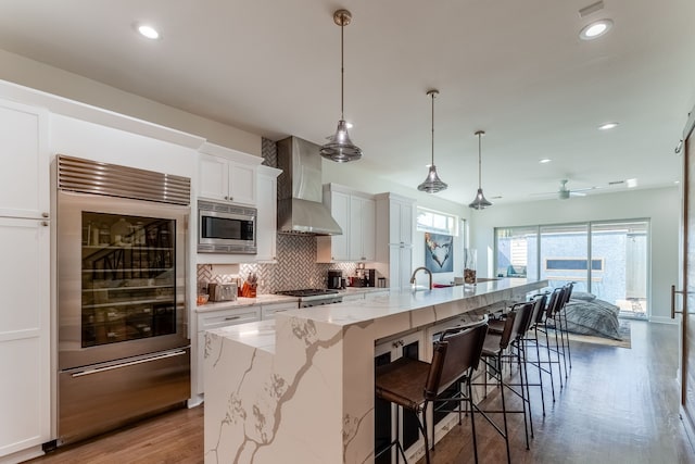 kitchen featuring hanging light fixtures, a spacious island, white cabinetry, wall chimney range hood, and built in appliances