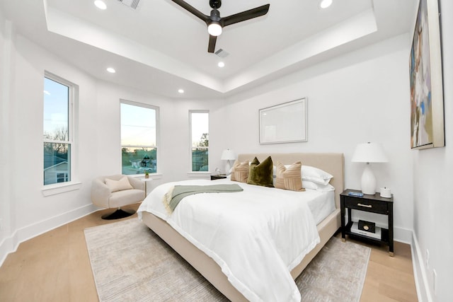 bedroom featuring ceiling fan, a raised ceiling, multiple windows, and light hardwood / wood-style flooring