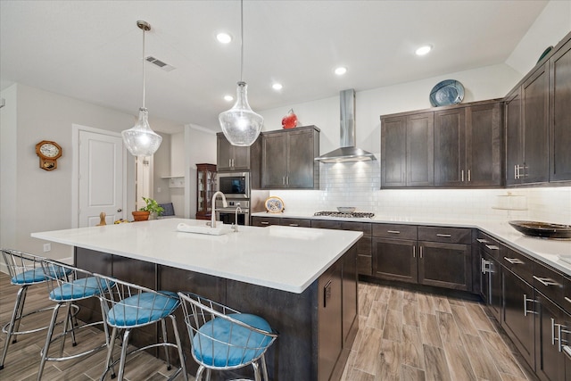 kitchen featuring appliances with stainless steel finishes, a kitchen breakfast bar, hanging light fixtures, a kitchen island with sink, and wall chimney range hood