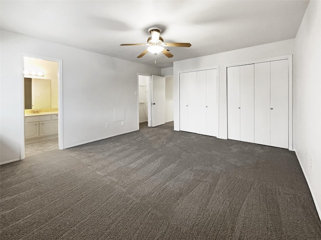 unfurnished bedroom featuring ensuite bathroom, ceiling fan, two closets, sink, and dark colored carpet