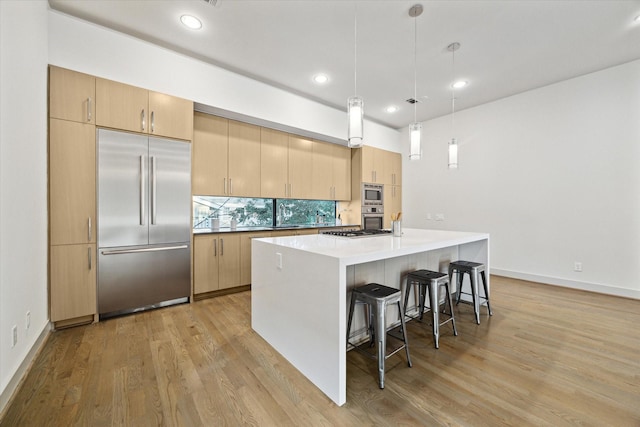 kitchen with light brown cabinets, built in appliances, hanging light fixtures, and an island with sink