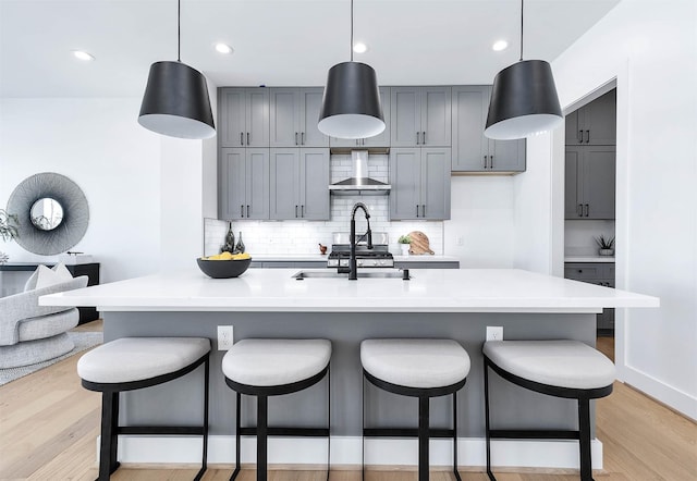 kitchen with hanging light fixtures, light hardwood / wood-style flooring, wall chimney range hood, and an island with sink