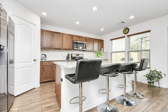 kitchen featuring appliances with stainless steel finishes, tasteful backsplash, light stone counters, a center island with sink, and a breakfast bar area