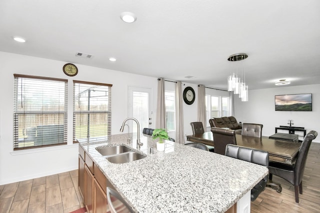 kitchen with dishwasher, hanging light fixtures, a kitchen island with sink, sink, and light stone counters