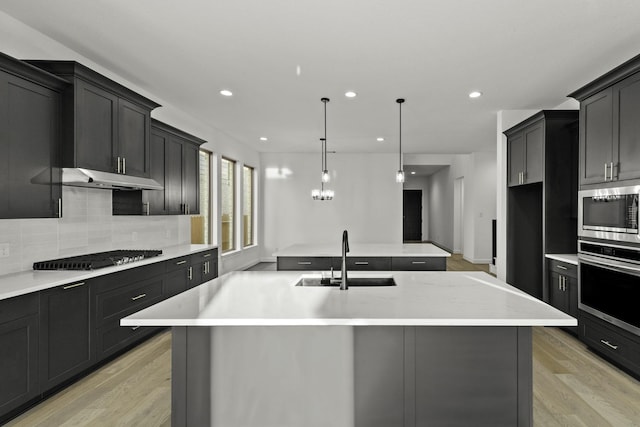 kitchen with under cabinet range hood, a sink, appliances with stainless steel finishes, backsplash, and an island with sink