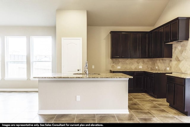 kitchen with a kitchen island with sink, lofted ceiling, light stone counters, dark brown cabinetry, and sink