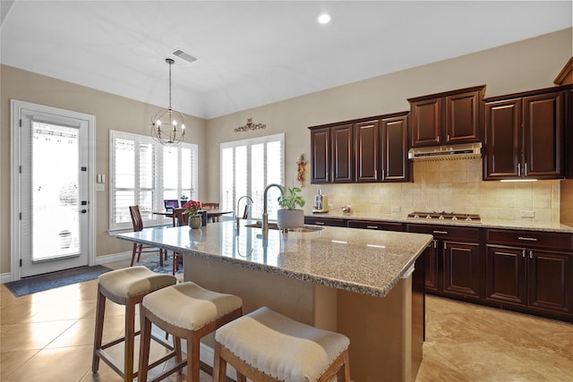 kitchen featuring stainless steel gas stovetop, an island with sink, decorative light fixtures, light stone counters, and sink